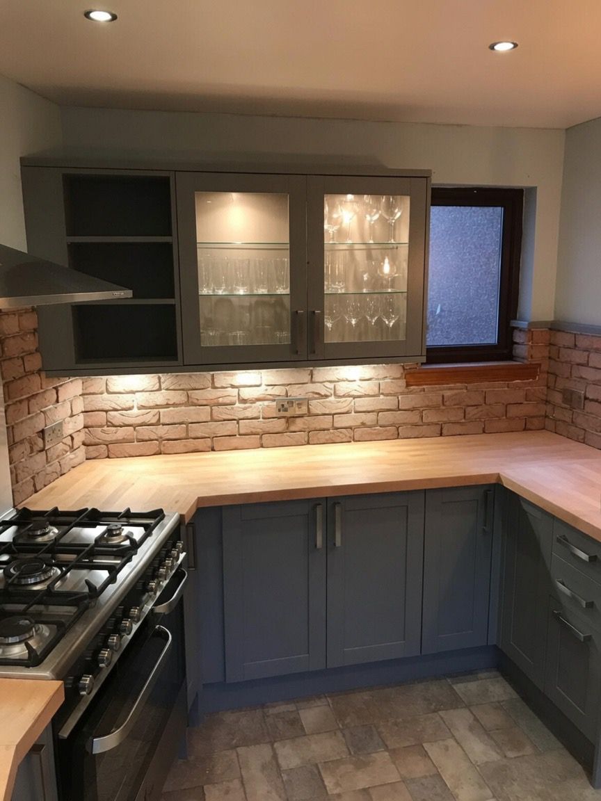 Grey shaker kitchen with an exposed brick splashback, solid timber worktops and glazed, lit display cabinets.